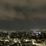 Jejak rudal melintasi langit malam di atas kota, terlihat cahaya ledakan di kejauhan selama serangan Iran-Israel