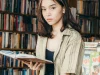 Seorang perempuan muda membaca buku di depan rak toko buku