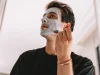 Seorang laki-laki mengoleskan masker perawatan pada wajahnya di depan cermin