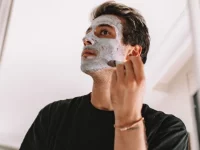 Seorang laki-laki mengoleskan masker perawatan pada wajahnya di depan cermin