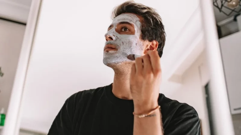 Seorang laki-laki mengoleskan masker perawatan pada wajahnya di depan cermin