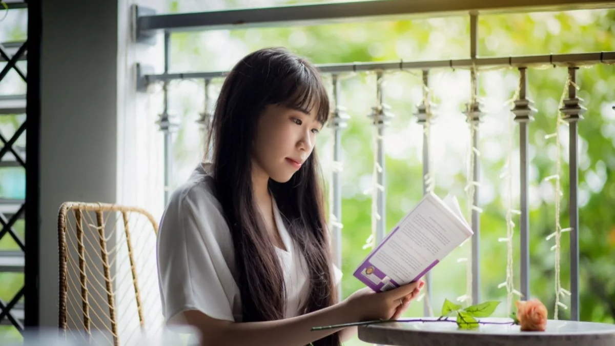 Seorang perempuan membaca buku di meja dekat jendela