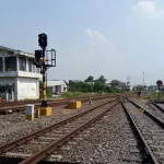 Bangunan rumah sinyal berdiri di tepi rel kereta api dengan tiang sinyal di sampingnya