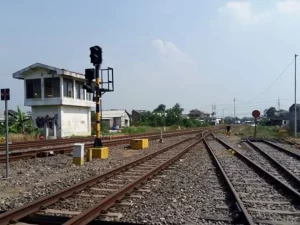 Bangunan rumah sinyal berdiri di tepi rel kereta api dengan tiang sinyal di sampingnya