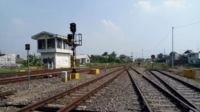 Bangunan rumah sinyal berdiri di tepi rel kereta api dengan tiang sinyal di sampingnya