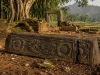 Ilustrasi ambang pintu batu andesit berhias relief sulur dan dua ukiran rosette, patah di salah satu ujungnya, tergeletak di antara reruntuhan bata berlumut dengan latar pohon beringin dan sawah