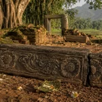 Ilustrasi ambang pintu batu andesit berhias relief sulur dan dua ukiran rosette, patah di salah satu ujungnya, tergeletak di antara reruntuhan bata berlumut dengan latar pohon beringin dan sawah