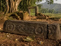 Ilustrasi ambang pintu batu andesit berhias relief sulur dan dua ukiran rosette, patah di salah satu ujungnya, tergeletak di antara reruntuhan bata berlumut dengan latar pohon beringin dan sawah
