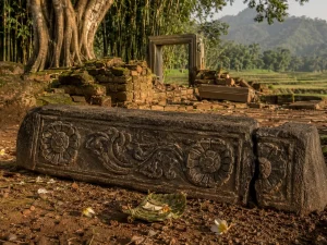 Ilustrasi ambang pintu batu andesit berhias relief sulur dan dua ukiran rosette, patah di salah satu ujungnya, tergeletak di antara reruntuhan bata berlumut dengan latar pohon beringin dan sawah