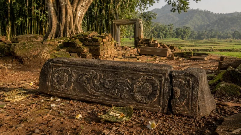 Ilustrasi ambang pintu batu andesit berhias relief sulur dan dua ukiran rosette, patah di salah satu ujungnya, tergeletak di antara reruntuhan bata berlumut dengan latar pohon beringin dan sawah