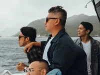 Sejumlah pria berdiri di geladak kapal dengan latar perbukitan dan laut