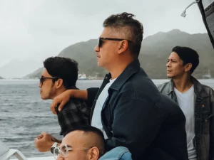 Sejumlah pria berdiri di geladak kapal dengan latar perbukitan dan laut