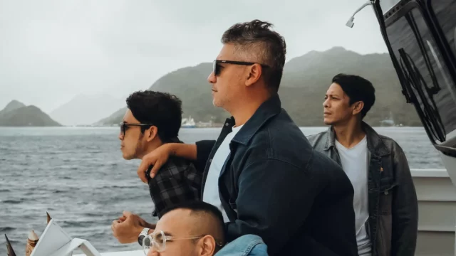 Sejumlah pria berdiri di geladak kapal dengan latar perbukitan dan laut