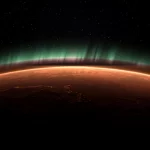 Ilustrasi lengkung cakrawala Mars berwarna jingga kemerahan dengan pijaran aurora hijau keunguan di atasnya dan latar angkasa berbintang
