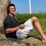 Baim Wong duduk bersandar di pohon dengan latar sawah dan tiang turbin angin di Jeneponto