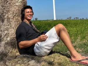 Baim Wong duduk bersandar di pohon dengan latar sawah dan tiang turbin angin di Jeneponto