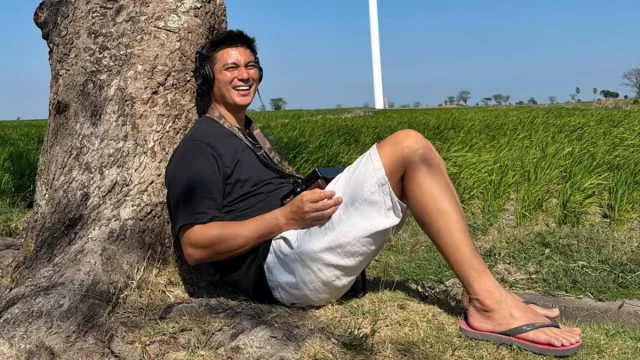 Baim Wong duduk bersandar di pohon dengan latar sawah dan tiang turbin angin di Jeneponto