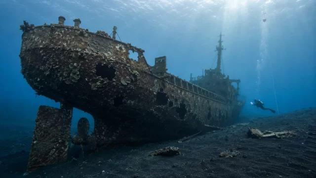 Ilustrasi bangkai kapal besi tua tergeletak di dasar laut berpasir gelap dengan lambung tertutup terumbu karang, lubang-lubang robekan pada dinding, kemudi dan baling-baling di buritan, serta seorang penyelam di kejauhan