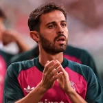Bernardo Silva mengenakan kaus pemanasan Real Madrid berwarna merah muda dan hijau sambil bertepuk tangan di tepi lapangan
