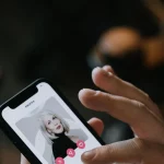 Tangan memegang ponsel pintar yang menampilkan kartu profil sebuah aplikasi kencan