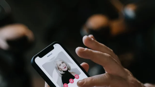 Tangan memegang ponsel pintar yang menampilkan kartu profil sebuah aplikasi kencan