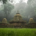 Kompleks stupa bata tua berlumut berdiri di padang rumput yang dikelilingi hutan tropis berkabut