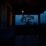 Ilustrasi beranda rumah kampung Sunda pada dini hari dengan satu bohlam teras menyala.