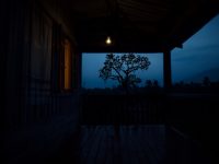 Ilustrasi beranda rumah kampung Sunda pada dini hari dengan satu bohlam teras menyala.