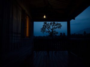 Ilustrasi beranda rumah kampung Sunda pada dini hari dengan satu bohlam teras menyala.