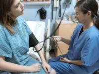 Petugas kesehatan berseragam biru memeriksa tekanan darah seorang pasien perempuan