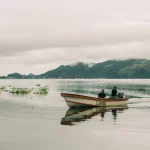 Dua orang menaiki perahu kayu bermesin di Danau Sentani dengan rumah panggung dan perbukitan berkabut di kejauhan