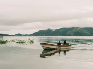 Dua orang menaiki perahu kayu bermesin di Danau Sentani dengan rumah panggung dan perbukitan berkabut di kejauhan