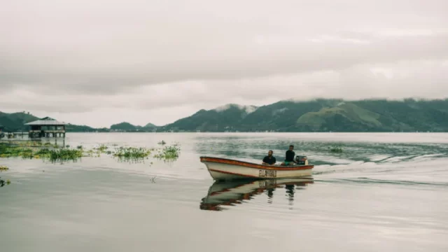 Dua orang menaiki perahu kayu bermesin di Danau Sentani dengan rumah panggung dan perbukitan berkabut di kejauhan
