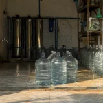Deretan galon bening dan tabung penyaring baja di sebuah depot air minum isi ulang.