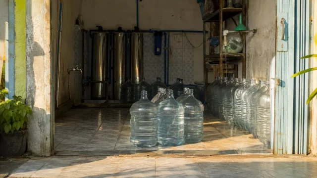 Deretan galon bening dan tabung penyaring baja di sebuah depot air minum isi ulang.