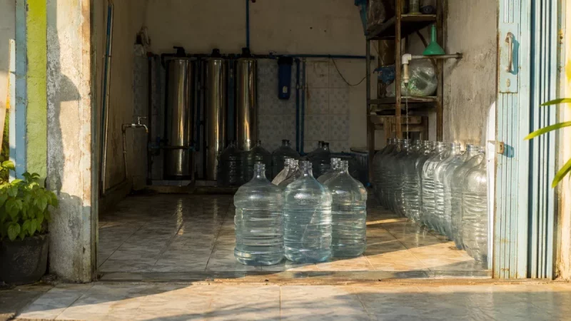 Deretan galon bening dan tabung penyaring baja di sebuah depot air minum isi ulang.