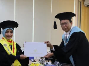 Seorang doktor baru bertoga menerima map penghargaan dari pimpinan sidang dalam ujian terbuka di ruang sidang kampus