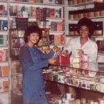Dua pekerja berdiri di balik etalase kaca sebuah toko buku dengan rak penuh buku dan barang dagangan, foto berstempel September 1982