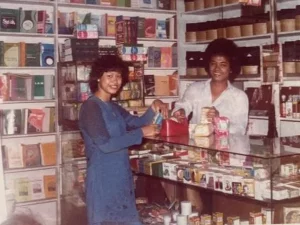 Dua pekerja berdiri di balik etalase kaca sebuah toko buku dengan rak penuh buku dan barang dagangan, foto berstempel September 1982