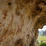 Ilustrasi dinding ceruk batu gamping berlapis kerak dengan gambar cadas berpigmen merah pudar bermotif figur manusia berkepala berjumbai, ikan, penyu, dan perahu, dengan lanskap karst serta tambak terlihat di kejauhan