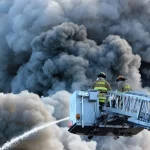 Dua petugas pemadam kebakaran di ujung tangga mobil damkar menyemprotkan air ke kepulan asap hitam