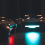 Dua stik pengendali konsol permainan dengan lampu biru menyala di atas permukaan gelap