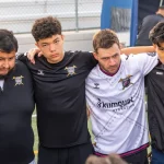 Ilustrasi sekelompok pemain sepak bola satu tim berangkulan membentuk lingkaran di pinggir lapangan bersama pelatihnya
