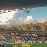 Stadion sepak bola dengan penonton di tribune pada sore hari berlatar perbukitan
