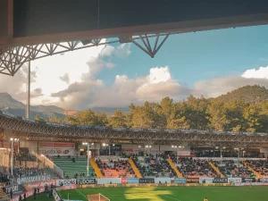 Stadion sepak bola dengan penonton di tribune pada sore hari berlatar perbukitan