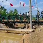 Jembatan rangka baja darurat terpasang di atas sungai, dengan jembatan lama yang ambruk terlihat di bawahnya