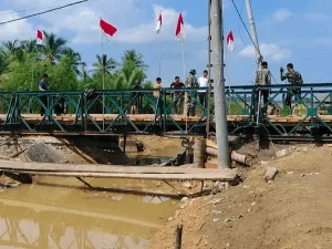 Jembatan rangka baja darurat terpasang di atas sungai, dengan jembatan lama yang ambruk terlihat di bawahnya