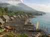 Ilustrasi kampung pesisir berumah panggung beratap daun di tepi pantai pasir gelap dengan perahu bercadik berlayar pandan yang ditambatkan, berlatar hutan sagu dan kerucut gunung berapi berasap
