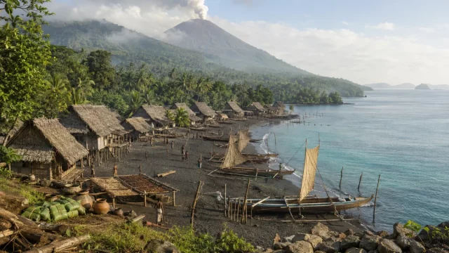 Ilustrasi kampung pesisir berumah panggung beratap daun di tepi pantai pasir gelap dengan perahu bercadik berlayar pandan yang ditambatkan, berlatar hutan sagu dan kerucut gunung berapi berasap