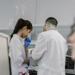 Dua peneliti berjas laboratorium bekerja di dekat instrumen, sementara seorang lainnya mencatat di depan komputer